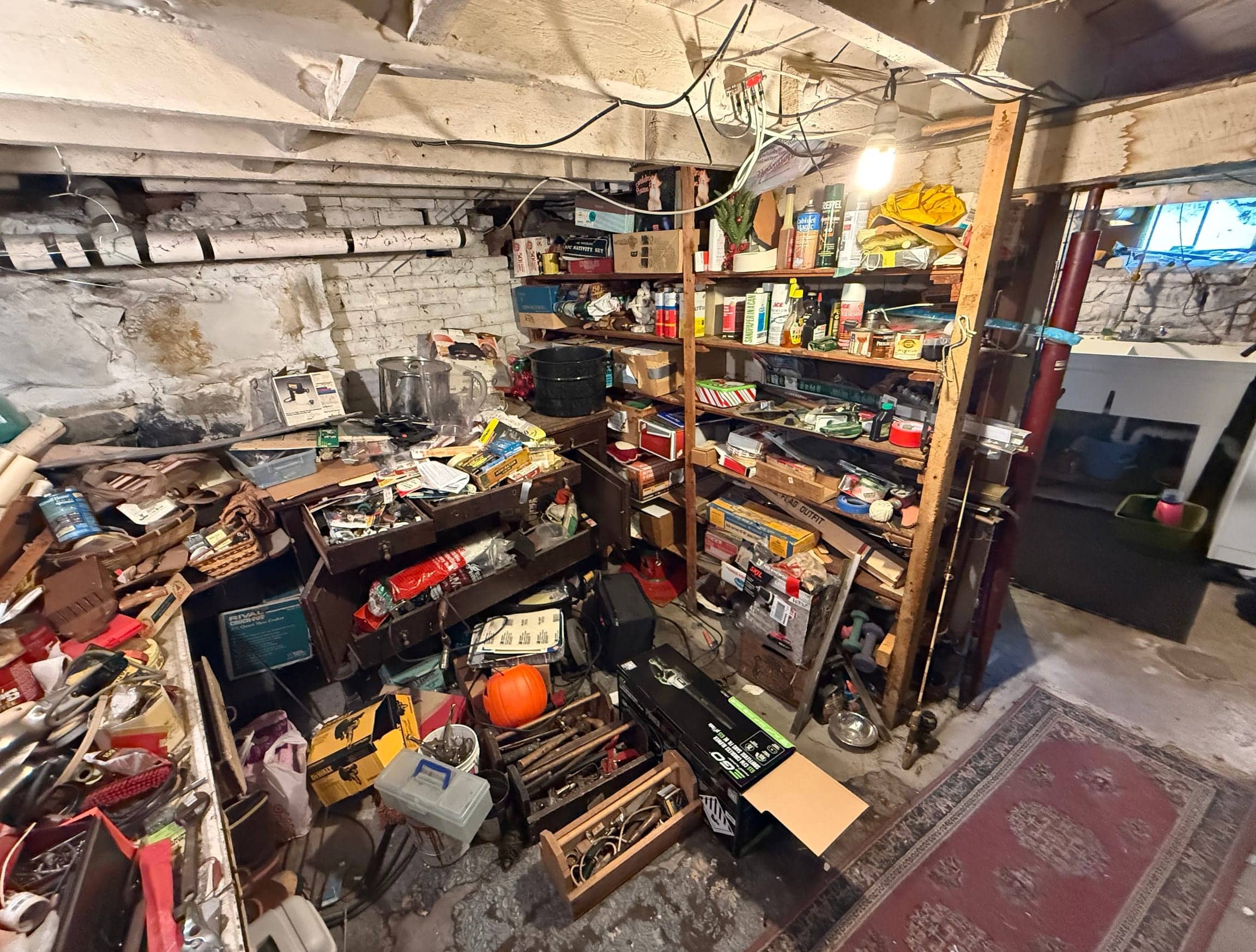 Cluttered basement with a packed workbench, shelves of paint cans and household chemicals, tools and boxes on the floor